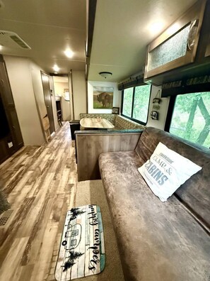 A compact RV interior with a kitchenette, a seating area with a cushion, and a window offering a view of trees.
