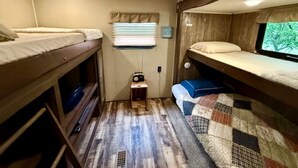 A compact RV interior with two bunk beds, a small kitchenette, and a window with blinds.