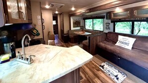 A compact RV interior with a kitchenette, a seating area, and a window providing a view of the outdoors. - Texas Hill Country Glamping Caravan for a Nature Escape near Hamilton Creek (Marble Falls)