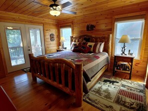 A wooden cabin bedroom with a bed, a ceiling fan, a window with a view, and a wooden floor.