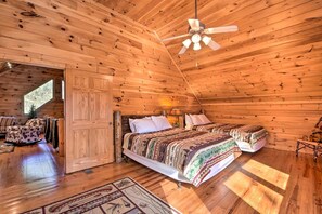 A log cabin bedroom with a ceiling fan, two beds, a rocking chair, and a large window.