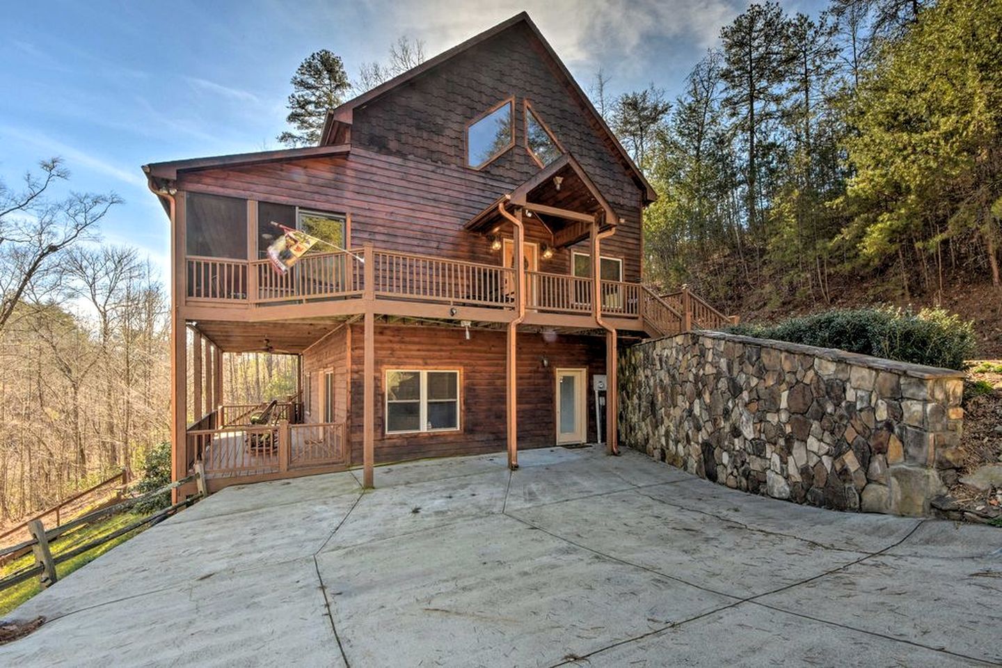 A two-story wooden cabin with a stone retaining wall, a balcony, and a large driveway.