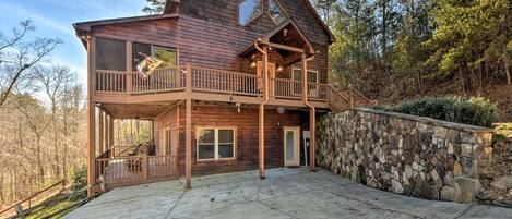 A two-story wooden cabin with a stone retaining wall, a balcony, and a large driveway.