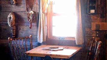 A rustic wooden interior with a dining table, four chairs, a hanging light fixture, and a window with curtains.