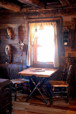 Un interior rústico de madera con una mesa de comedor, cuatro sillas, una lámpara colgante y una ventana con cortinas.
