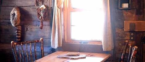 A rustic wooden interior with a dining table, four chairs, a hanging light fixture, and a window with curtains.