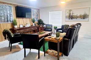Un salon comprenant une table à manger en bois, des chaises noires, un canapé, un téléviseur fixé au mur et deux plantes en pot.