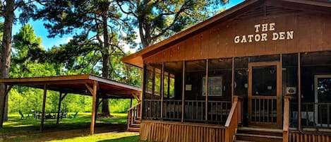 A wooden building labeled 'The Gator Den' with a covered outdoor seating area and a staircase leading to the entrance.