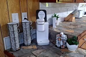 A kitchen with a coffee maker, stacked mugs, a sign, and a plant.