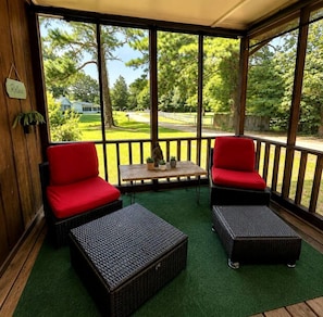 A covered outdoor seating area with two red cushioned chairs and two wicker ottomans, a small table with potted plants, and a view of a green landscape with trees and a pathway.