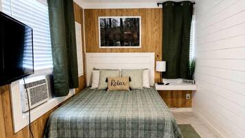 A bedroom with a bed, a ceiling fan, a window with blinds, a wall-mounted TV, and a bedside table with a lamp.
