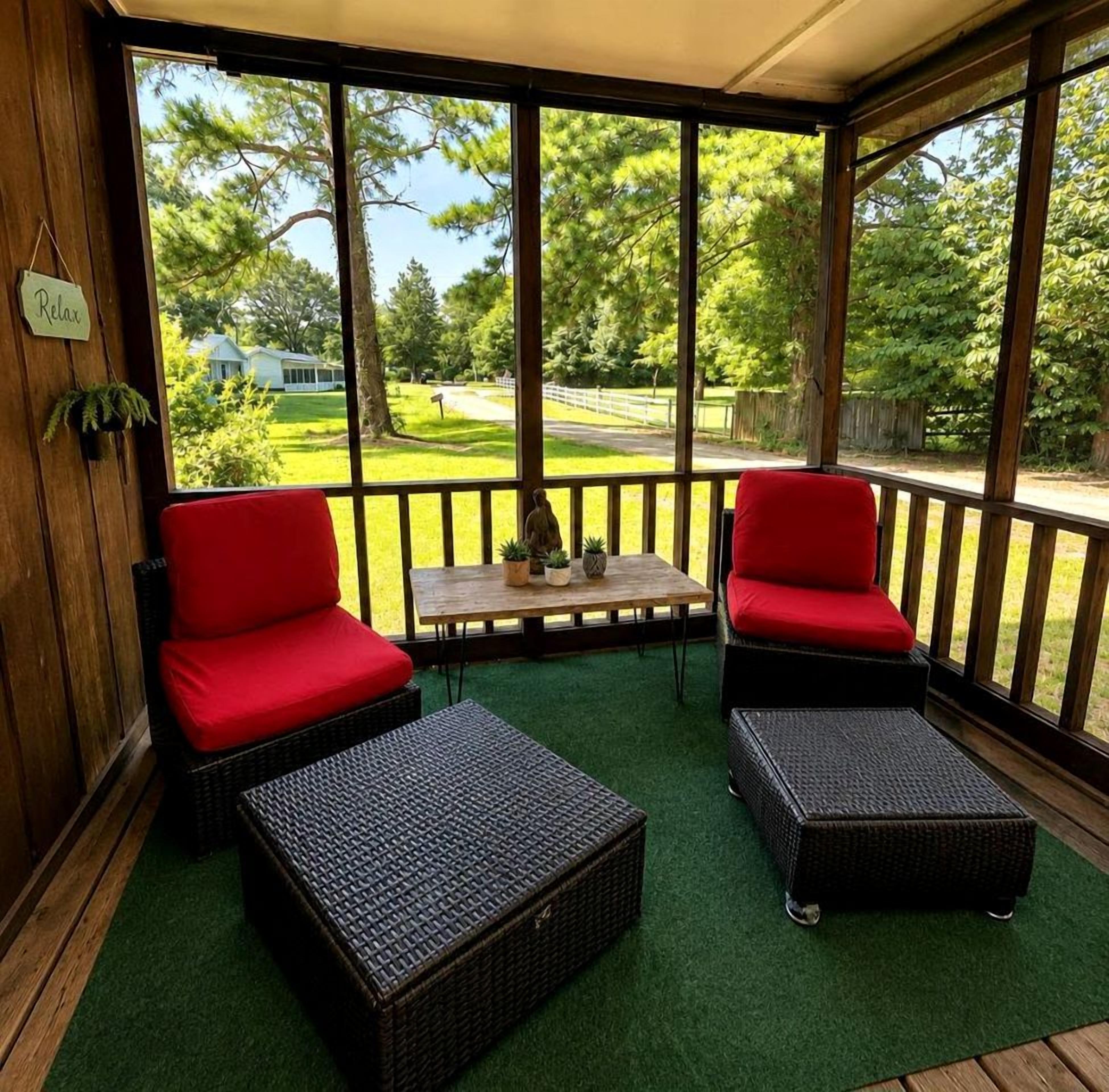 Un espace extérieur aménagé, abrité et doté de deux fauteuils rouges rembourrés et de deux poufs en osier, ainsi qu’une petite table ornée de plantes en pot, le tout offrant une vue sur un paysage verdoyant, avec des arbres et un sentier.