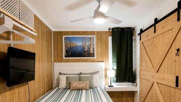 A bedroom with a bed, a mounted TV, a ceiling fan, a window with curtains, and a wooden door.