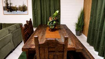 Une table de salle à manger en bois avec des bancs, une plante verte en guise de centre de table, un tableau encadré au mur et un rideau vert.