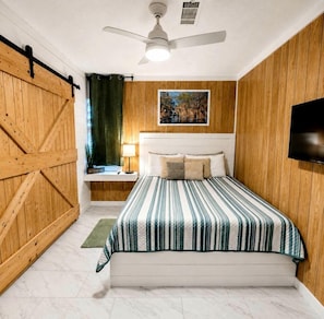 Een slaapkamer met een bed, een plafondventilator, een wandgemonteerde televisie, een houten wandpaneel en een raam met gordijn.