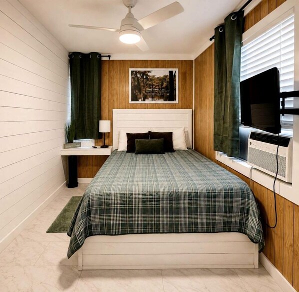 Ein Schlafzimmer mit einem Bett, einem Deckenventilator, einem an der Wand montierten Fernseher und einem Fenster mit Vorhängen.