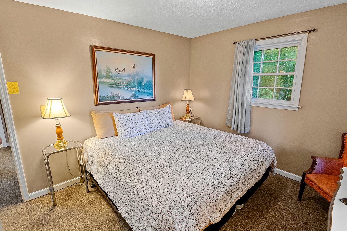 A bedroom with a bed, two bedside lamps, a painting, a window with curtains, and a chair.