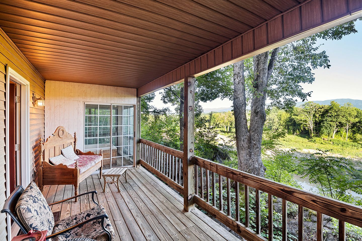 A wooden deck with a bed and chairs, a glass door, and a view of a forest and river.