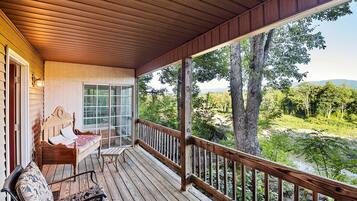 A wooden deck with a bed and chairs, a glass door, and a view of a forest and river.