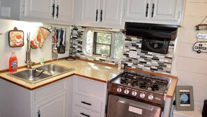 A compact kitchen with white cabinets, a stainless steel sink, a stove, and a window with a view. - Texas Hill Country Vintage Caravan Glamping (Marble Falls)