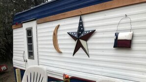 A wooden deck with two white plastic chairs with red cushions, a small round table, and a white mobile home with a blue exterior. - Texas Hill Country Vintage Caravan Glamping (Marble Falls)
