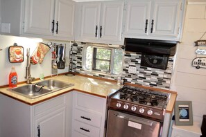 A compact kitchen with white cabinets, a stainless steel sink, a stove, and a window with a view. - Texas Hill Country Vintage Caravan Glamping (Marble Falls)