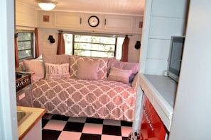 A compact RV interior with a kitchenette, a bed with patterned bedding, and a window with curtains. - Texas Hill Country Vintage Caravan Glamping (Marble Falls)