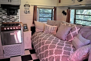A cozy RV interior with a kitchenette, a bed with patterned bedding, and a window with curtains. - Texas Hill Country Vintage Caravan Glamping (Marble Falls)