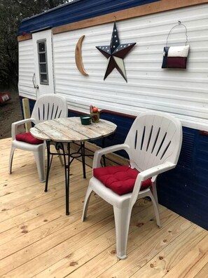 A wooden deck with two white plastic chairs with red cushions, a small round table, and a white mobile home with a blue exterior.