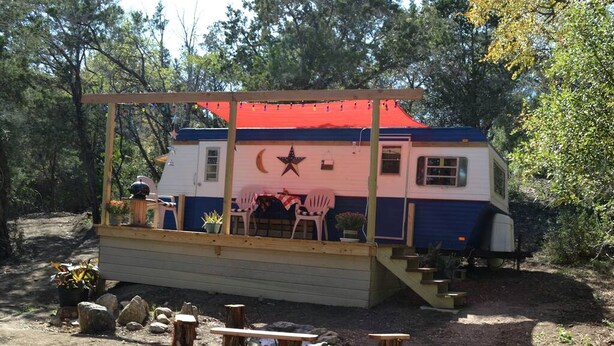 A small cabin with a porch, a table, and chairs, surrounded by trees. - Texas Hill Country Vintage Caravan Glamping (Marble Falls)