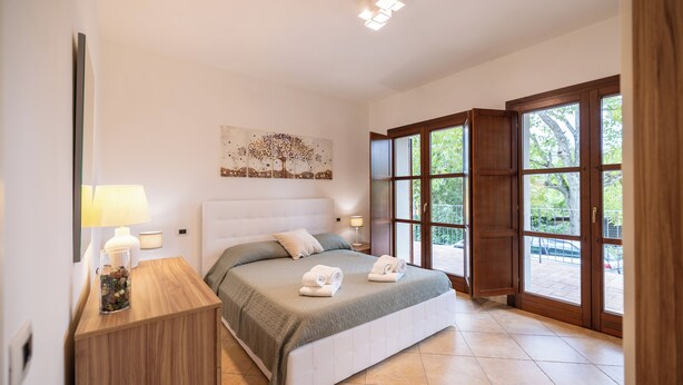 Une chambre avec un grand lit, une table de chevet, une lampe, un tableau et une vue sur l’extérieur grâce à une porte vitrée.
