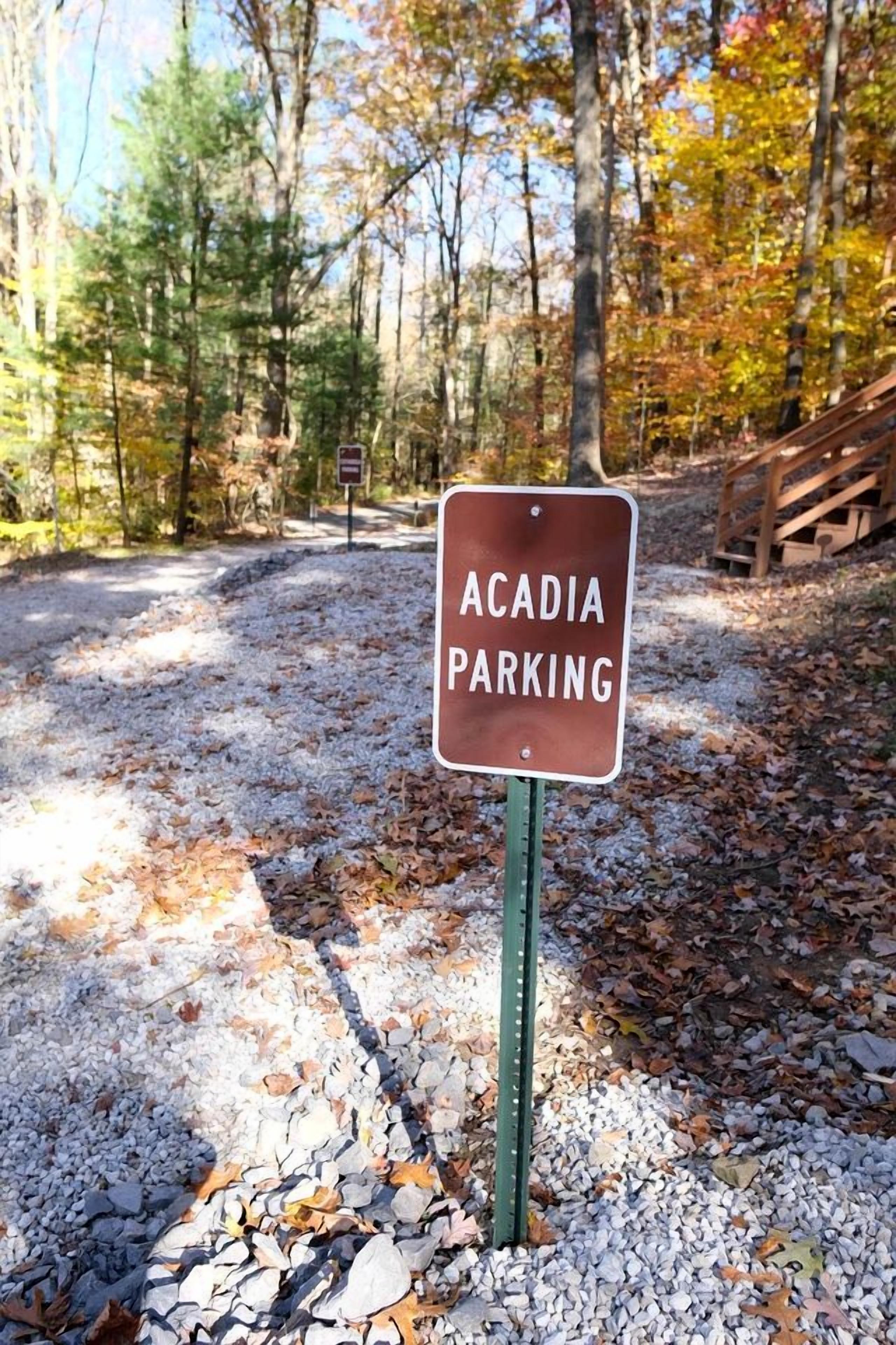 A sign for Acadia parking in a forested area with gravel paths and a wooden bridge.