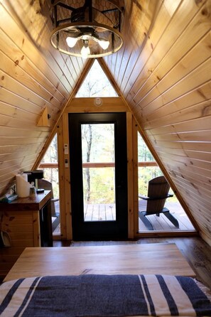 Una cabaña de madera con techo triangular, un escritorio con lámpara, una silla y vista al exterior por una puerta.