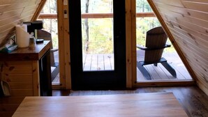 Una cabaña de madera con techo triangular, un escritorio con lámpara, una silla y vista al exterior por una puerta.