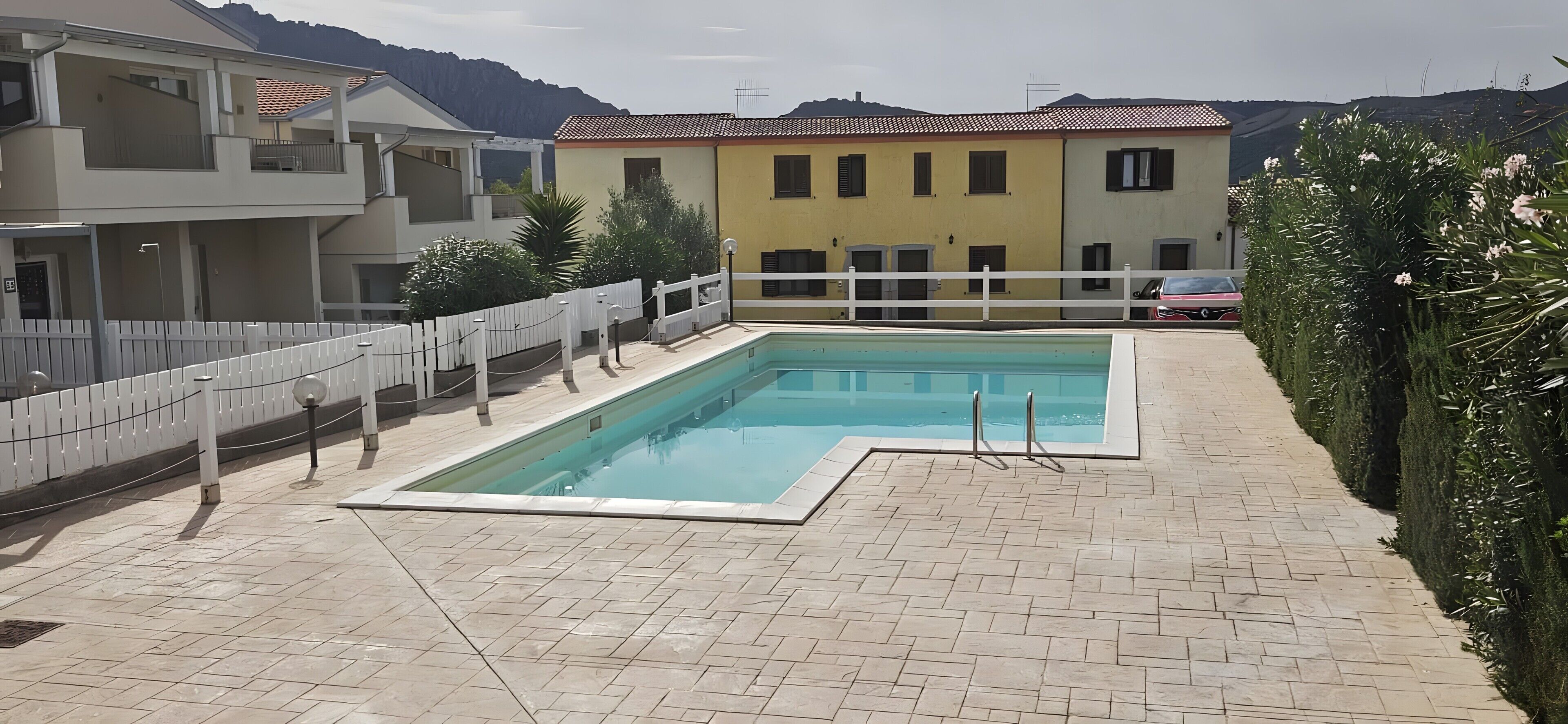 Una piscina rectangular con una solana blanca, rodeada por una valla blanca y macetas con plantas.