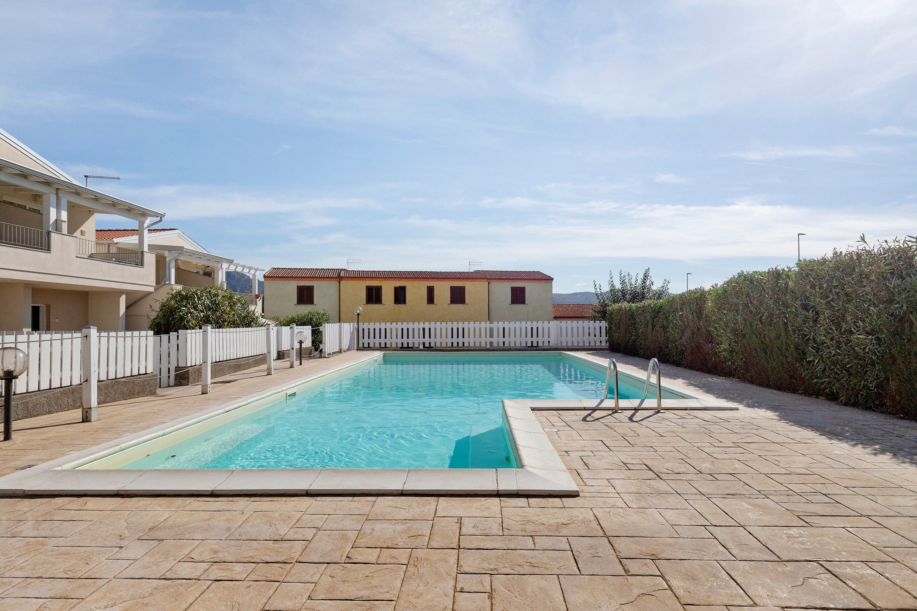 A rectangular swimming pool with a white fence surrounding it, set against a backdrop of buildings and a clear sky.