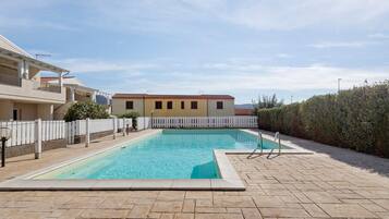 A rectangular swimming pool with a white fence surrounding it, set against a backdrop of buildings and a clear sky.