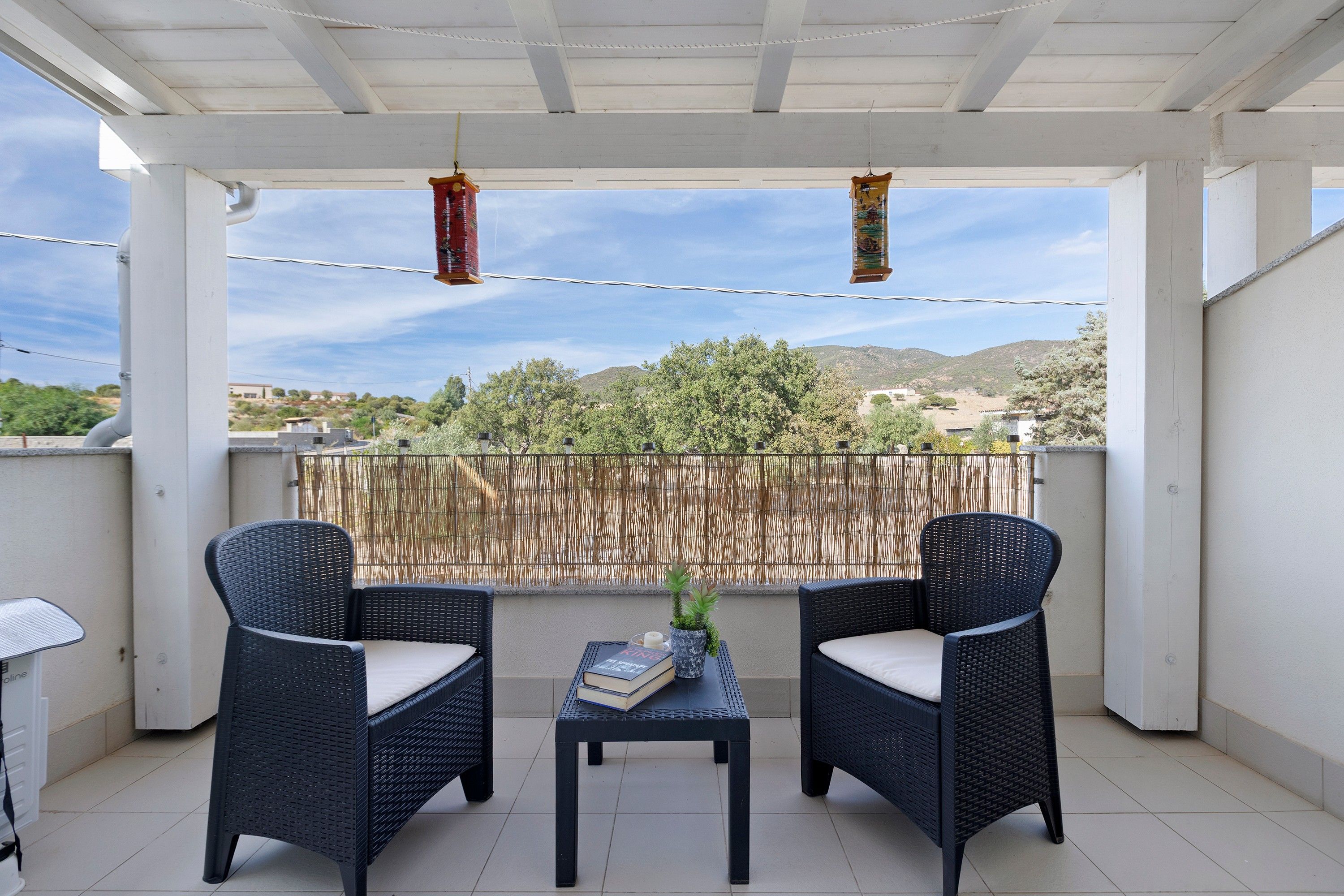 A covered balcony with two wicker chairs, a small table, and a view of a mountainous landscape.