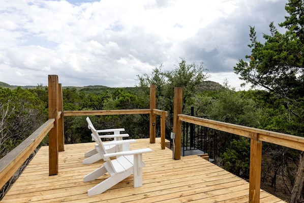 Una terraza de madera con barandilla, dos sillas Adirondack blancas y vistas a un área boscosa.