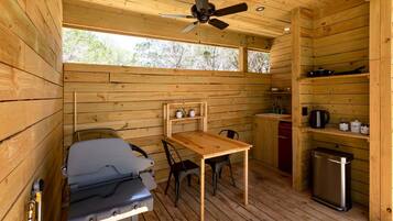 A compact cabin with a wooden interior, a ceiling fan, a table with chairs, a kitchenette, and a small trash bin.