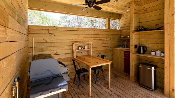 A compact cabin with a wooden interior, a ceiling fan, a table with chairs, a kitchenette, and a small trash bin. - Peaceful Romantic Yurt with Hot Tub in Wimberley, Texas (Wimberley)