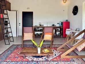 Un salon avec une table basse en bois, deux fauteuils rembourrés, un tapis à motifs, une petite plante et, au fond, un coin cuisine.