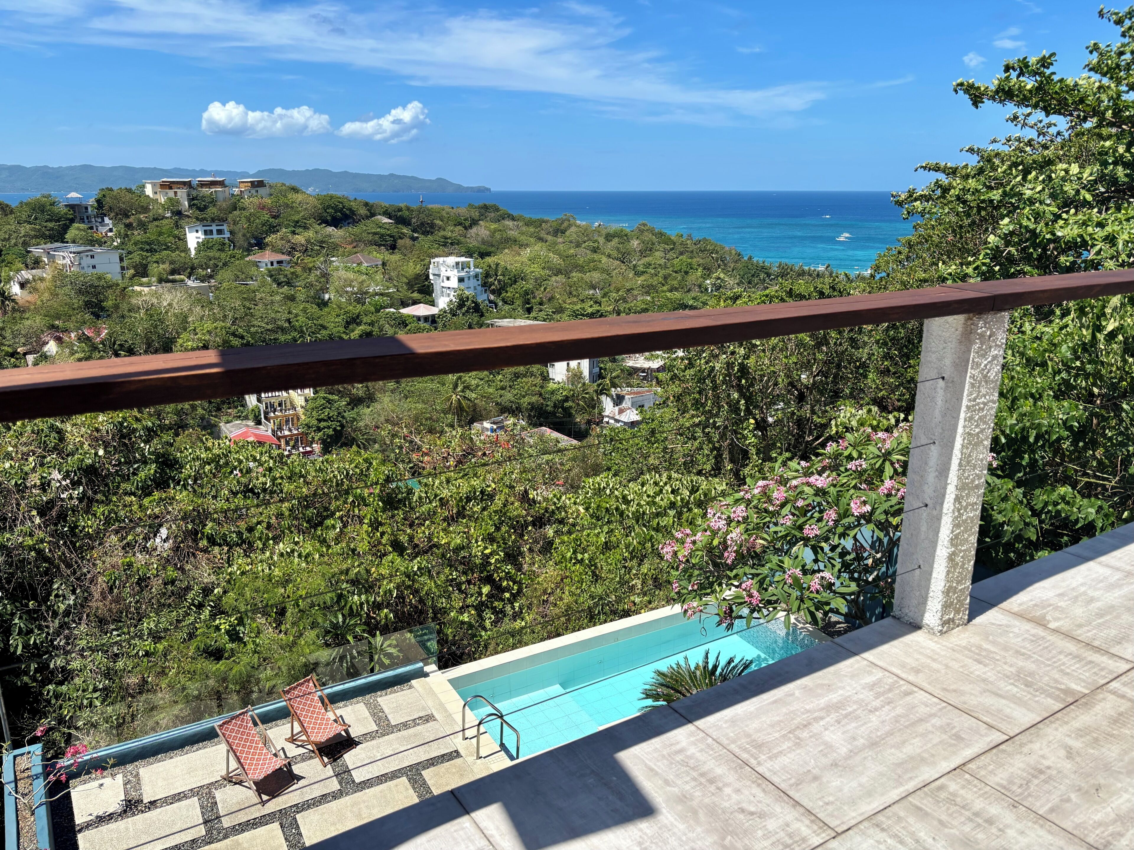 A balcony with a pool, overlooking a coastal area with houses and greenery.