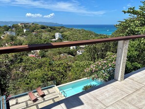 A balcony with a pool, overlooking a coastal area with houses and greenery.
