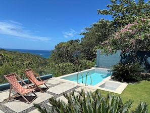 A pool with two lounge chairs, surrounded by greenery and a view of the ocean.