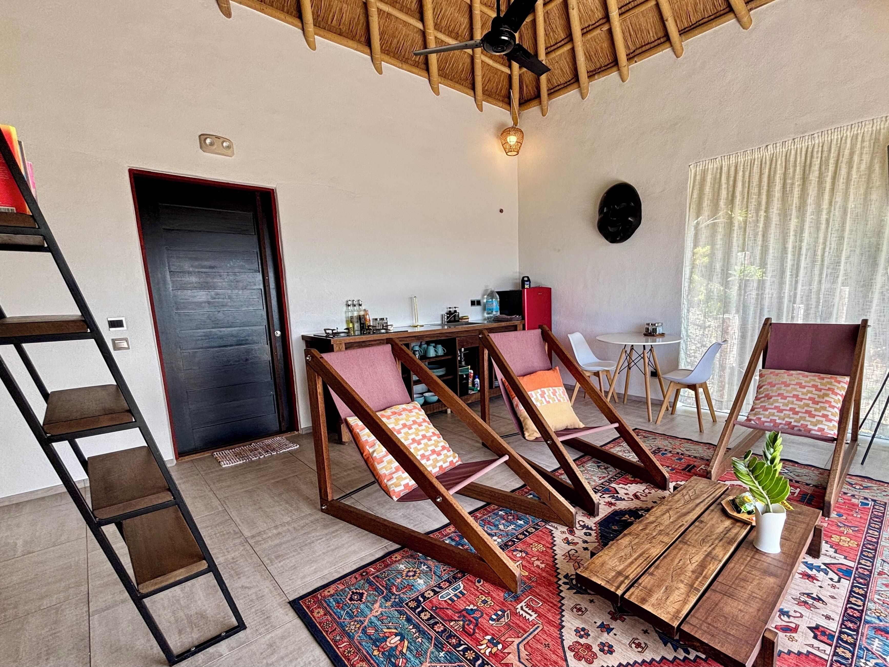 A living room with a wooden ceiling, a black door, a dining table, two wooden chairs, a small table with a plant, and a colorful rug.