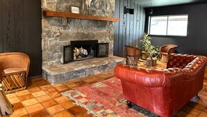 A stone fireplace with a mantel shelf, a red leather sofa, a wooden chair, and a patterned rug in a room with wooden floors and a window.