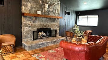 A stone fireplace with a mantel shelf, a red leather sofa, a wooden chair, and a patterned rug in a room with wooden floors and a window.