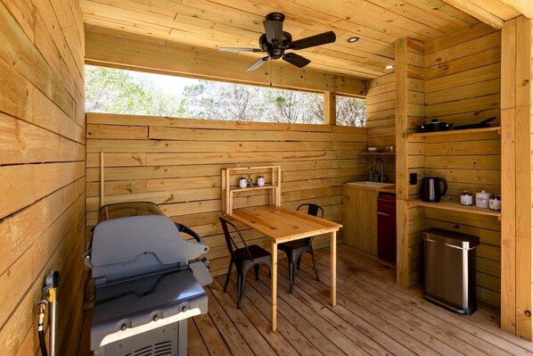 A compact cabin with a wooden interior, a ceiling fan, a table with chairs, a kitchenette, and a small trash bin.