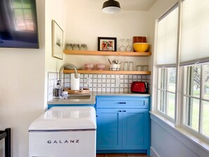 A kitchen with a white Galanz refrigerator, blue cabinets, a red toaster, and a window with blinds.